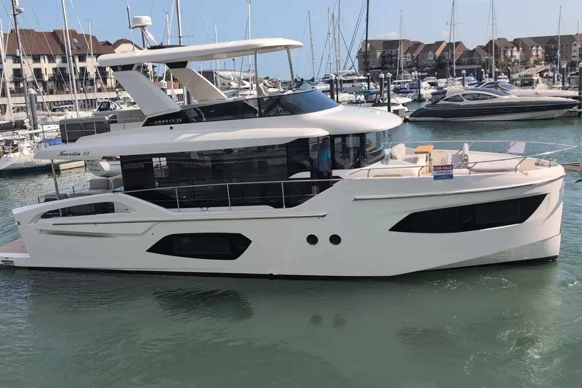 A sleek white Navetta 53 yacht docked in a marina, with a "For Sale" sign visible on the deck.